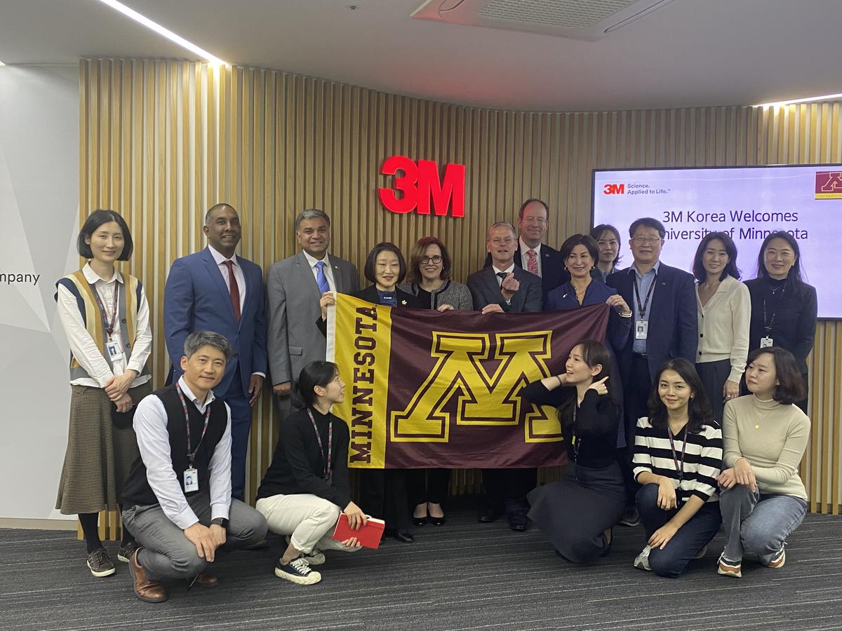 Group photo of 17 people in front of 3M logo, two women in the center of the frame are holding a UMN flag